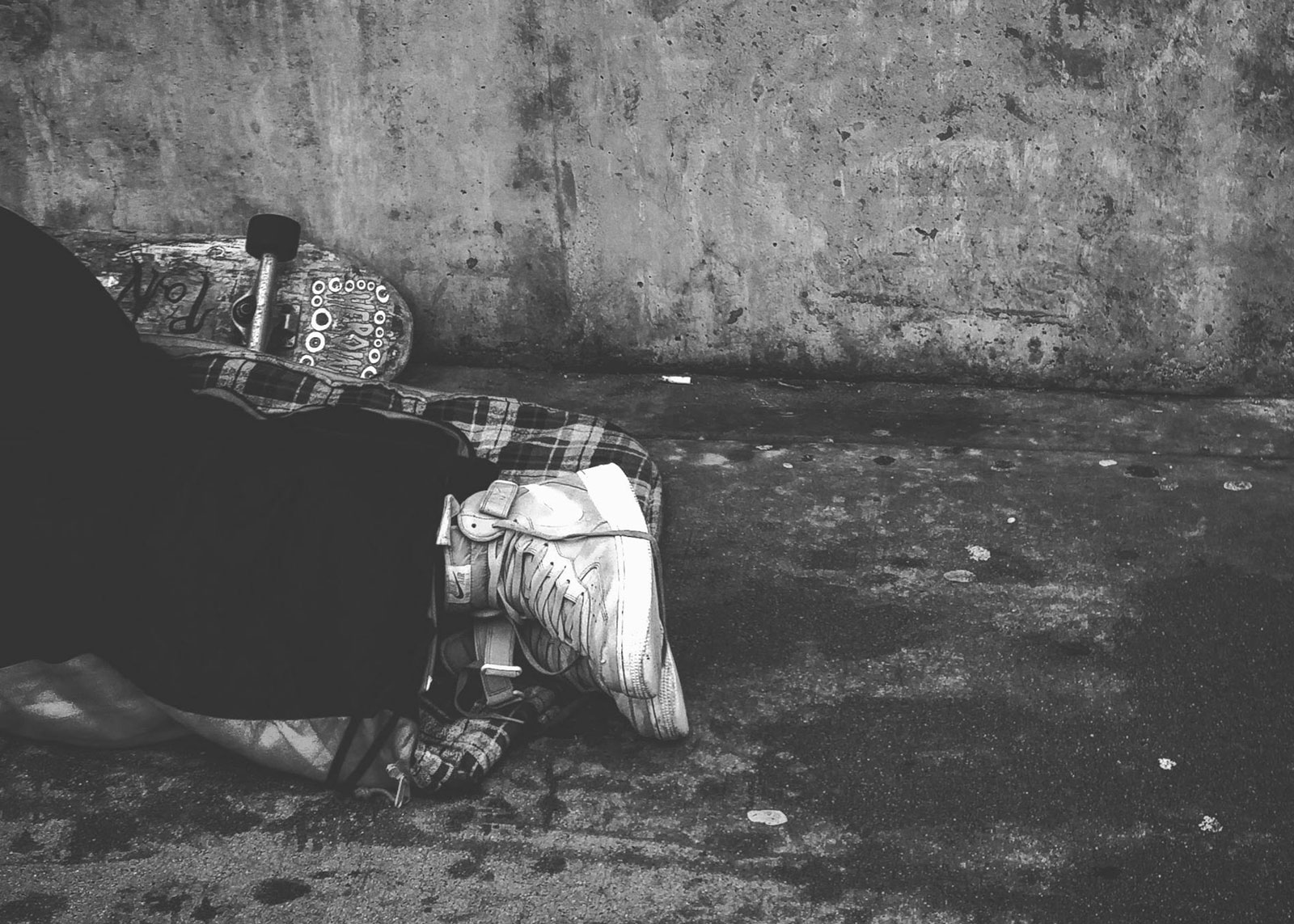 Black and white image of a person sleeping on the ground, with their legs and feet visible. A skateboard is placed nearby, subtly hinting at homelessness, reflecting the challenges of mental and behavioral health in San Francisco.